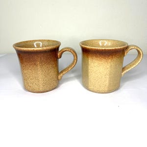 Puede incluir: Dos tazas de cerámica con exterior moteado color beige y borde marrón. Las tazas tienen un asa curvada y una parte superior ligeramente ensanchada. Las tazas están vacías y se encuentran sobre una superficie blanca.