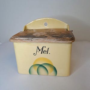 Può includere: Un contenitore di stoccaggio a parete in ceramica giallo vintage con un coperchio in legno. Il contenitore ha un design a sole e cerchi verdi e la parola "Mel." scritta in nero.