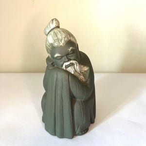 Puede incluir: Una figura de cerámica gris oscuro que representa a una persona sentada con un peinado de moño y una barba larga. La figura tiene detalles en plata metálica en el cabello, las cejas y la barba, y está en una pose de descanso con los brazos cruzados.