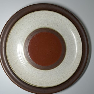 Puede incluir: Un plato de cerámica marrón con un centro blanco y un círculo rojo en el medio. El plato tiene un esmalte moteado.