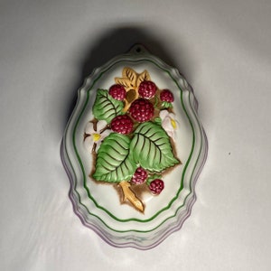Puede incluir: Molde de cerámica blanca con un diseño en relieve de hojas verdes, una rama marrón y frambuesas rojas. El molde tiene un borde morado y verde.