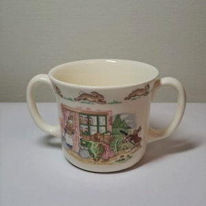 May include: A white ceramic mug with two handles features a colourful illustration of bunnies shopping at a store. The mug has a cream-coloured background with a green, pink, and brown design.