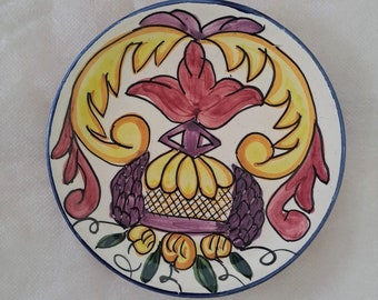 May include: A white ceramic plate with a blue rim features a hand-painted design of a stylized floral motif in shades of yellow, red, purple, and green.