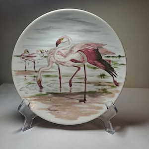 May include: A white ceramic plate featuring a painted image of pink flamingos standing in a shallow body of water. The plate has a brown rim and the word "Flamingos" is written in black at the bottom.