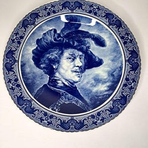 Può includere: Un piatto decorativo blu e bianco con un ritratto di un uomo con un cappello piumato. Il ritratto è circondato da un bordo dettagliato con motivi floreali e geometrici. Un'opera d'arte classica.