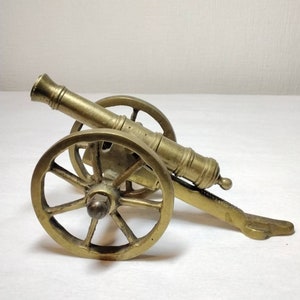 May include: A small, antique brass cannon with spoked wheels. The miniature artillery piece features a detailed barrel, carriage, and a lever. The cannon is a decorative item, likely a collectible or display piece.