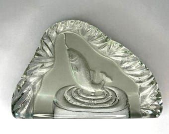 May include: A clear, triangular glass dish with an etched design of a fish leaping from water. The fish is in the center, surrounded by concentric circles. The edges of the dish have a textured, leaf-like pattern.