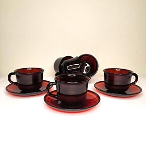 Può includere: Un set di sei tazze da tè e piattini in vetro rosso scuro. Le tazze hanno una forma arrotondata con un manico e i piattini sono circolari. Le tazze e i piattini sono disposti su una superficie bianca.