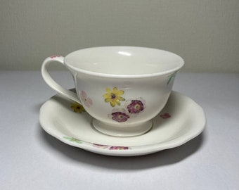 May include: A white teacup and saucer set with a floral pattern. The cup has a handle and a wide, rounded bowl. The saucer has a scalloped edge.