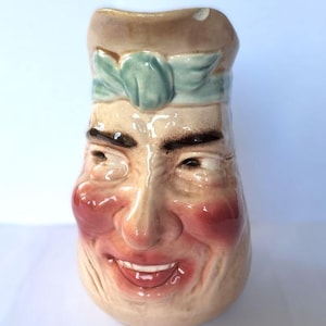 Peut inclure: Un pichet en céramique en forme de visage souriant avec un chapeau bleu et vert. Le pichet a une peau de couleur brun clair avec des joues roses et des yeux bruns.