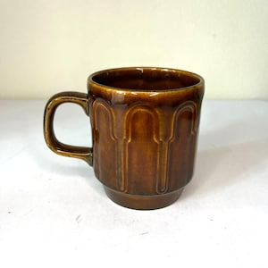 May include: A brown ceramic mug with a handle. The mug has a glossy finish and a textured, vertical grooved design. The mug is a warm brown color and appears to be vintage.
