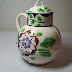 May include: A white ceramic pitcher with a lid, decorated with green leaves and red and purple flowers. The pitcher has a round body and a handle.