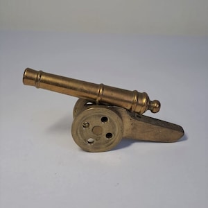 May include: A small, brass cannon with a wheel on one side. The cannon is about 4 inches long and is made of solid brass.
