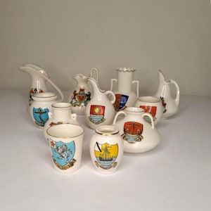 Puede incluir: Una colección de ocho jarrones y jarras de cerámica en miniatura con varios diseños. Cada pieza presenta un escudo de armas diferente con detalles coloridos. Los jarrones son blancos con detalles dorados.
