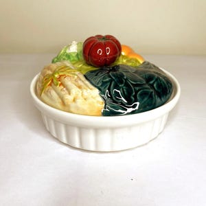 Puede incluir: Una fuente de horno de cerámica blanca con una tapa esculpida decorada con diversas verduras coloridas, incluyendo un tomate rojo, hojas de color verde oscuro y tallos claros, sobre una base redonda con bordes acanalados.