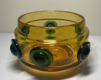 May include: A round amber glass bowl with a textured surface and four green and blue glass circles around the rim.