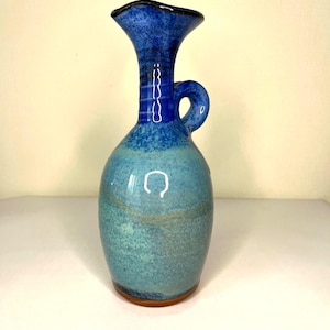 Può includere: Un vaso in ceramica fatto a mano con una glassa blu e turchese. Il vaso presenta un bordo svasato, un piccolo manico e una superficie strutturata. La base è di colore marrone scuro. Questo vaso decorativo è adatto per esporre fiori o come opera d'arte a sé stante.