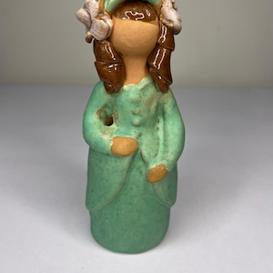 May include: A ceramic figurine of a woman in a green dress and hat, with brown hair and floral accents. The figurine has a light brown face and hands, and the dress has button details. The overall design is simple and rustic.