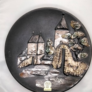 Puede incluir: Un plato de cerámica negro con un diseño en relieve de un pueblo con edificios y un árbol. El plato tiene un esmalte de color plateado y una etiqueta que dice "Ruscha Art echt Handwerk".