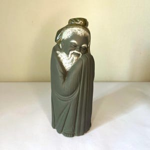 May include: A matte dark gray ceramic figurine of an elderly person with a long white beard and hair, wearing a traditional robe. The figure has closed eyes and is depicted in a peaceful, resting posture with hands tucked into the garment.