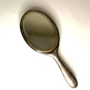 May include: Oval-shaped, silver-toned hand mirror. The mirror has a reflective surface and a handle for easy use. The frame and handle show signs of wear, adding to its vintage appeal. A classic beauty accessory.