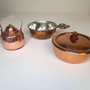 May include: Three miniature copper kitchenware items: a small kettle with a handle, a bowl with a handle, and a lidded pot. The kettle and pot have a shiny finish, while the bowl shows signs of wear. The items are arranged on a white surface.