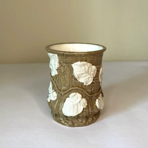 Peut inclure: Un récipient cylindrique en céramique avec un extérieur brun texturé imitant l'écorce d'un arbre. La surface est ornée d'appliques de feuilles blanches en relief et de fins détails de vignes entrelacées. L'intérieur du récipient est blanc.