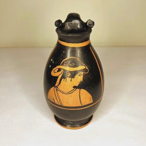 Puede incluir: Jarrón de cerámica negra con una ilustración dorada de una persona con sombrero y toga. El jarrón tiene una banda dorada alrededor del cuello y una parte superior decorativa. Un ejemplo clásico de cerámica griega antigua.