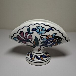 May include: A white ceramic napkin holder with a blue, green, orange, and pink floral design. The holder has a fan-shaped top and a round base.