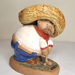 May include: A ceramic figurine of a man wearing a large sombrero, white shirt, red scarf, and blue pants. The figure is bent over, holding a small object. The base is oval-shaped and has a painted grassy area.