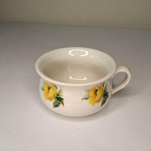 Könnte beinhalten: Weiße Suppenschüssel aus Keramik mit gelbem Rosenmuster. Die Schüssel hat eine abgerundete Form und einen Henkel.