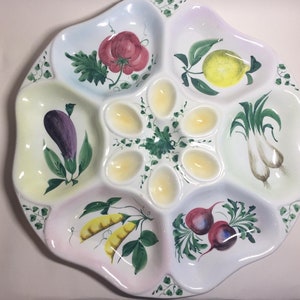 May include: A white ceramic serving dish with six sections. The centre section has six indentations for eggs. The other sections are decorated with painted vegetables, including a tomato, lemon, aubergine, peas, and radishes.