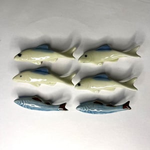 Può includere: Sei statuette di pesci in ceramica. Due file di tre pesci ciascuna. La fila superiore ha pesci biancastri con pinne blu. La fila inferiore ha pesci blu con dettagli marroni. I pesci sono disposti su una superficie bianca.