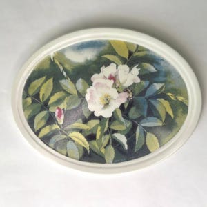 Puede incluir: Arte floral enmarcado en blanco de forma ovalada. La obra presenta una pintura a la acuarela de rosas blancas y rosas con hojas y capullos verdes. El fondo es una mezcla de tonos verdes y azules.