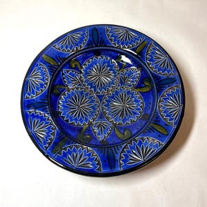 Puede incluir: Un plato de cerámica azul con un diseño decorativo de flores estilizadas en blanco y verde. El plato tiene un borde azul oscuro y un acabado brillante.