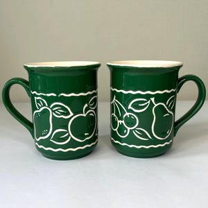 Puede incluir: Dos tazas de cerámica verde con diseños de frutas blancas. Las tazas tienen un borde blanco y un interior blanco.