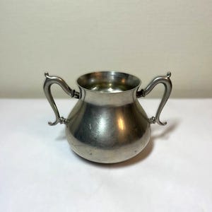 May include: A silver creamer with two handles. The creamer is made of pewter and has a rounded body.