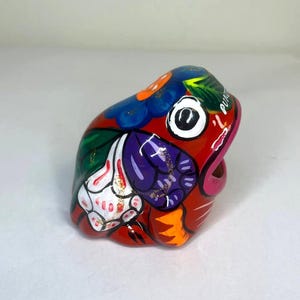 以下が含まれることがあります： 赤、青、緑、紫、オレンジ、白など、鮮やかな色で手描きされたセラミックのカエルの置物です。カエルは口を大きく開けており、花柄で飾られています。カエルには「PUEBLA」というラベルが貼られています。