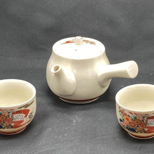 Könnte beinhalten: Eine weiße Teekanne aus Keramik mit Blumenmuster und zwei passende Teetassen. Die Teekanne hat einen Henkel und einen Ausguss. Die Teetassen haben ein Blumenmuster an der Seite.