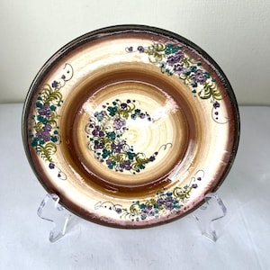 Może przedstawiać: Ozdobny ceramiczny talerz z brązowym i kremowym środkiem, ozdobiony kwiatowymi wzorami w kolorze fioletowym, zielonym i kremowym. Talerz ma ciemnobrązową obwódkę i jest prezentowany na przezroczystym stojaku.