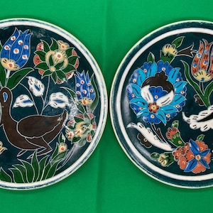 Puede incluir: Dos platos redondos de cerámica con fondo azul verdoso y diseños florales blancos, azules, rojos y amarillos. Un plato presenta un pájaro marrón de cuello largo y un diseño floral. El otro plato presenta un diseño floral con una gran flor azul.