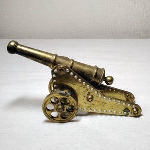 May include: A brass miniature cannon with two wheels, a barrel, and decorative rivets. The cannon is a golden color and sits on a white surface. The cannon is a decorative item.