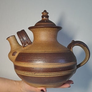 Può includere: Una teiera in ceramica marrone e beige con un coperchio e un design a strisce. La teiera ha un corpo arrotondato e un beccuccio laterale.