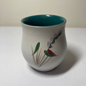 May include: A white ceramic mug with a green rim and a floral design featuring a red, white, and green flower.