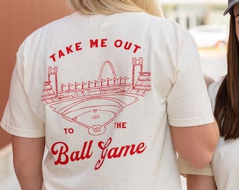 May include: Two women wearing white t-shirts with red baseball-themed graphics. The woman on the left is wearing a blue denim hat with the letters "STL" embroidered on it. The woman on the right is wearing a gray baseball cap with the letters "STL" embroidered on it. The woman on the left's shirt says "Take me out to the ball game" and features a drawing of a baseball stadium. The woman on the right's shirt says "Saint Louis" and features a baseball and a baseball glove.