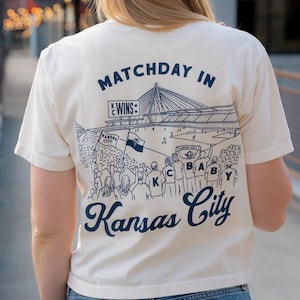 May include: Cream-colored t-shirt with a navy blue graphic of a stadium scene and the text "Matchday in Kansas City." The design includes a bridge, fans, and the words "KC" and "Wins."