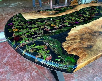 Peut inclure: Une grande table ovale avec un plateau en résine époxy noire et un piétement en bois naturel. Le plateau en résine présente un motif de fleurs et de feuilles pressées dans des tons de vert, rouge et jaune.