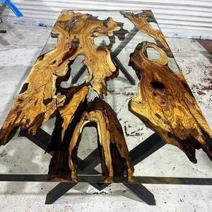 May include: A large wooden table top with a natural wood grain pattern and black epoxy resin filling in the gaps. The table top is covered with a clear glass top. The table has a black metal base with a cross-shaped design.