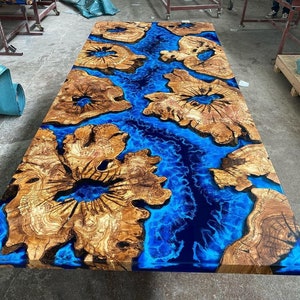 Puede incluir: Una gran tapa de mesa de madera con una incrustación de resina epoxi azul. La resina tiene un patrón de remolino que se asemeja al agua. La madera es de color marrón claro con un patrón de veta natural.