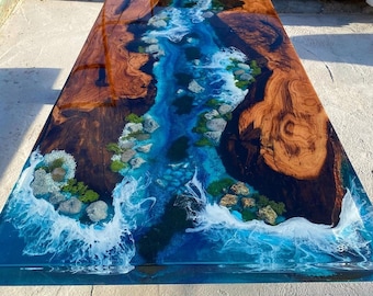May include: A large wooden table with a blue and white epoxy resin river design. The resin features white waves and green moss-like accents. The wood is a dark brown with a natural grain pattern.